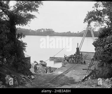 [Mango [Mago], The Lagoon], Burton Brothers Studio, Fotostudio, Juli 1884, Dunedin, Schwarz-Weiß-Fotografie, in der Mitte befindet sich eine Gruppe fidschianischer Männer am Fuße eines Holzkran, der sich auf einem Steg befindet. Eine Eisenbahnlinie fährt vom Steg hoch, wo zwei lange Boote festgemacht werden. Links stehen zwei Menschen unter einem Schirm. Auf beiden Seiten im Vordergrund stehen Bäume auf Felswänden. Die Lagune ist im Hintergrund, mit hohem Land mit Vegetation bedeckt Stockfoto
