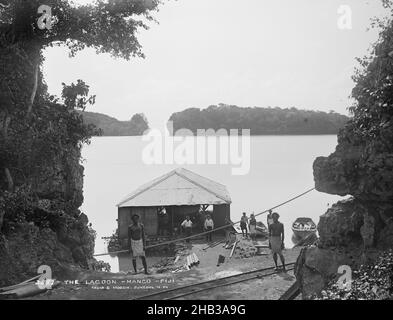 The Lagoon, Mango [Mago], Fidschi, Burton Brothers Studio, Fotostudio, Juli 1884, Dunedin, Schwarz-Weiß-Fotografie, Bootsschuppen am Ufer der Lagune, mit Eisenbahnlinie vor der Lagune. Im Hintergrund sind zwei Landmassen zu sehen. Zwei lange Boote befinden sich neben dem Bootshaus, drei Männer stehen direkt vor dem Bootshaus und drei zu seiner Rechten. Zwei Fidschi-Männer stehen in der Nähe des Zentrums bergauf und nahe dem Vordergrund. Im Vordergrund auf beiden Seiten befinden sich Bäume und rechts eine Felswand Stockfoto