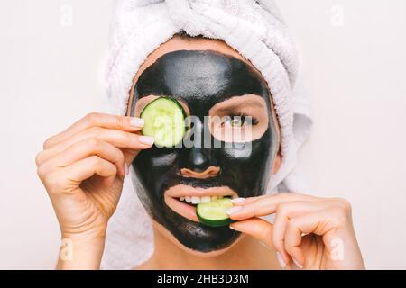 Schöne Frau, die schwarze reinigende Holzkohle-Gesichtsmaske mit Gurkenscheiben anwendet. Gesichtshautpflege Stockfoto