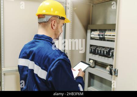 Elektriker in Schutzanzug und Helm mit Tablet und versuchen, Fehler zu beheben, während sie neben dem Armaturenbrett in Schwerindustrie-Werk stehen. Stockfoto