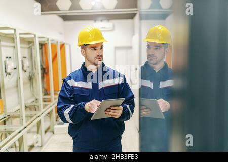 Elektriker in Uniform und Helm auf dem Kopf mit Tablet und Überprüfung auf dem Armaturenbrett in der Leitwarte in der Schwerindustrie. Stockfoto