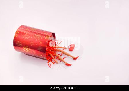 Eine rot-goldene umgestürzte Geschenkbox mit roten glänzenden Zinnmuscheln und Frauenarmen und -Beinen mit roten Schuhen, die auf weißem Hintergrund herausfallen. Eine rote Handtasche besi Stockfoto