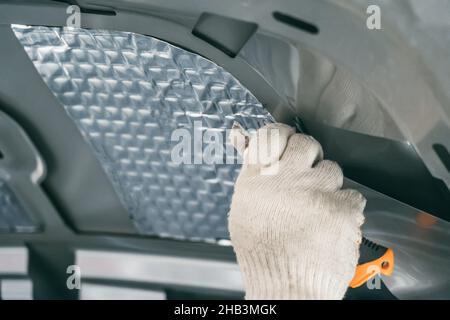 Arbeiter klebt Schalldämmmaterial an der Innenseite der Motorhaube. Prozess der Installation der Schalldämmung im Auto. Stockfoto