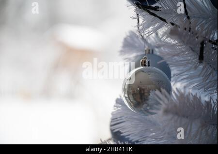 Nahaufnahme einer blauen Weihnachtskugel aus Glas, die auf einem weißen weihnachtsbaum mit schneebedeckter Natur im Hintergrund hängt. Stockfoto