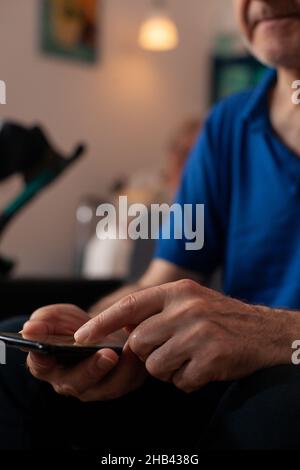 Nahaufnahme von unkenntlich alten Händen, die das Smartphone zu Hause auf dem Sofa halten. Älterer pensionierter Mann berührt Gerät mit moderner Technologie, während er auf der Couch im Wohnzimmer sitzt. Person, die Gadget verwendet. Stockfoto