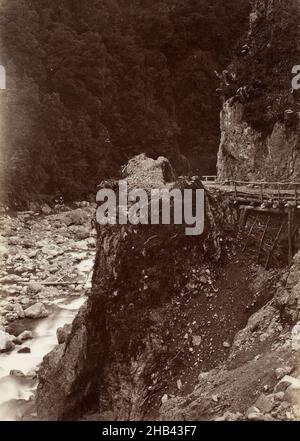 Otira Gorge, West Coast Road, Burton Brothers Studio, Fotostudio, Dunedin, Schwarzweiß-Fotografie, Blick auf einen Teil der Landschaft der Otira-Schlucht. Flusswasser kann entlang der nach links gerichteten Seite des Bildes gesehen werden, und eine bebaute Straße ist auf der nach rechts gerichteten Seite zu sehen. Der Hügel im Hintergrund ist mit einheimischen Sträuchern und Bäumen verkleidet, während die Uferlandschaft auf Bodenhöhe von einer Kombination aus Felsen und Steinen bedeckt ist Stockfoto