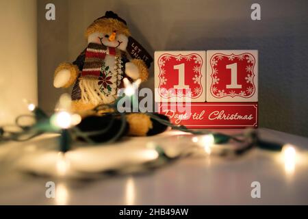 Elf Tage bis Weihnachten Countdown-Kalender aus Holzblöcken auf einem Mantel mit Lichtern und einem Plüsch Spielzeug Schneemann. Stockfoto