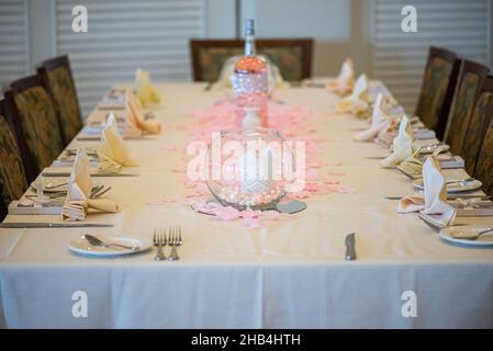 Dinner-Setting mit elegantem Glas-Kerzenhalter im Mittelpunkt mit weißer Kerze und pink/weißen Perlen im Inneren und umgeben von rosa Blütenpedalen Stockfoto