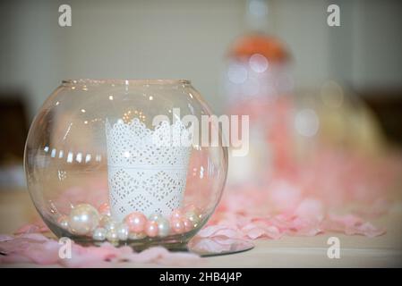 Eleganter Kerzenhalter aus Glas mit weißer Kerze und pink/weißen Perlen im Inneren und umgeben von rosa Blumenpedalen am Hochzeitstisch Stockfoto