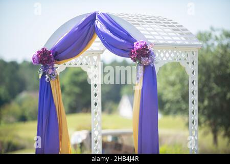 Majestätischer weißer Hochzeitssbogen, der in violettes und gelbes Tuch gehaucht und bei der Hochzeitszeremonie im Freien mit roten Blumen geschmückt ist Stockfoto