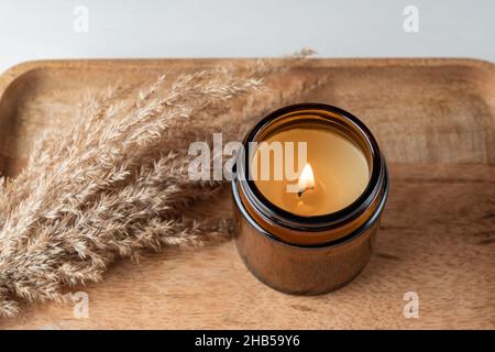 Handgemachte Kerze aus Paraffin und Sojawachs im Glasgefäß mit trockenem Schilfgras auf Holztablett. Kerzenherstellung Stockfoto