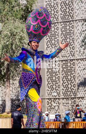 Dubai, VAE, 09.12.2021. Lächelnder Mann auf Stelzen, der farbenfrohe Kleidung der Expo 2020 und einen riesigen Rundhut während der Expo 2020 Dubai Daily Parade trug. Stockfoto