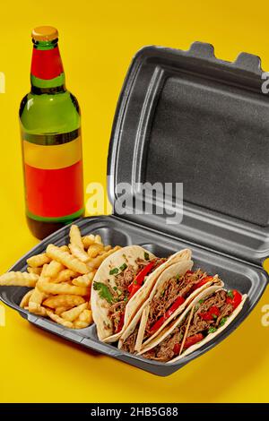 Birria Tacos im Takeaway-Behälter mit Pommes Frites und einer Flasche Getränk auf gelbem Hintergrund Stockfoto