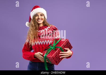 Glückliche junge Frau trägt Weihnachtsmann Hut hält rote Geschenkbox auf lila Hintergrund. Weihnachtsfeier Stockfoto