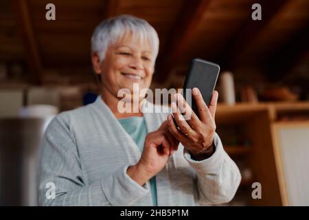 Lächelnde multikulturelle ältere Frau, die eine Nachricht auf dem Smartphone liest, während sie in der modernen Küche steht. Stockfoto