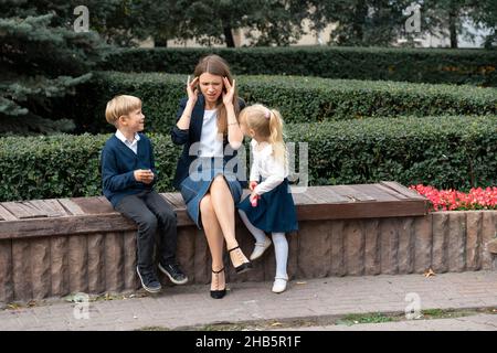 Müde Mutter, die Probleme mit frechen lauten Kindern, berühren Stirn, leiden unter Kopfschmerzen, schwierigst Kind, Kinder-Tantrum und Manipulation Co Stockfoto