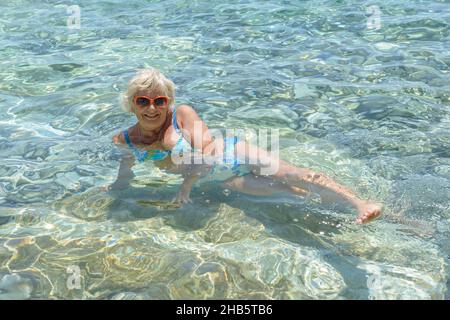Lächelnde ältere Frau liegt im klaren, transparenten Meerwasser eines flachen Küstenstrandes von Lucice, Stockfoto