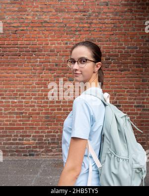 Porträt einer Schülerin in einem T-Shirt und mit einem blauen Rucksack auf dem Hintergrund einer braunen Ziegelwand, Kopierraum. Tourismuskonzept für Studenten. Fremd e Stockfoto