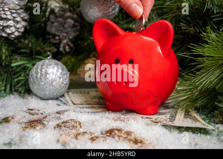 Hand, die eine Münze in ein rotes Sparschwein auf Goldmünzen und Papiergeld bedeckt mit Schnee und Weihnachten grünen Tannenzweigen mit Kugeln verziert. Savin Stockfoto