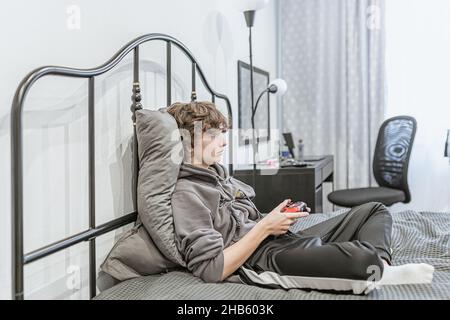 Teenager-Mädchen mit kurzen braunen lockigen Haaren, bekleidet mit ungepflegten Heimkleidung, sitzt auf einem ungefertigten Bett und spielt auf der Computerkonsole. Leidenschaft für Computer Stockfoto