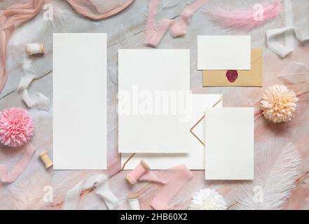 Leere Karten und Umschläge zwischen Pastellblumen, Seidenbändern und Federn auf Marmor-Draufsicht. Romantische Szene mit Hochzeitssuite Mockups flat lay, p Stockfoto