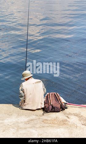 Ein eingeflügeliger Fischer in einem Panamahut sitzt auf einem betonierten Pier mit einer Angelrute am Wasser. Stockfoto