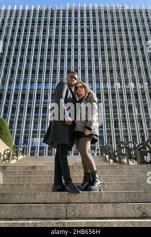 Von unten stilvolles multirassisches Paar in stilvoller Oberbekleidung, das sich an sonnigen Tagen in der Stadt auf den Stufen vor dem modernen Wolkenkratzer umarmt und steht Stockfoto