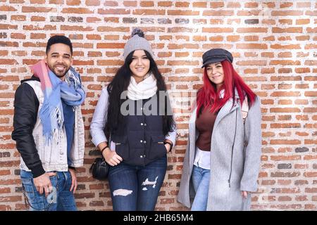 Positive multirassische junge Freundinnen und Freundinnen tragen trendige Casual-Outfits, die in der Nähe einer Ziegelsteinmauer stehen und vor der Kamera lächeln Stockfoto