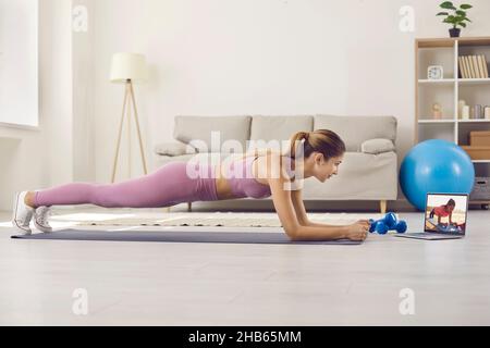 Die junge, sportliche Frau arbeitet zu Hause und macht eine Planke vor ihrem Laptop. Stockfoto