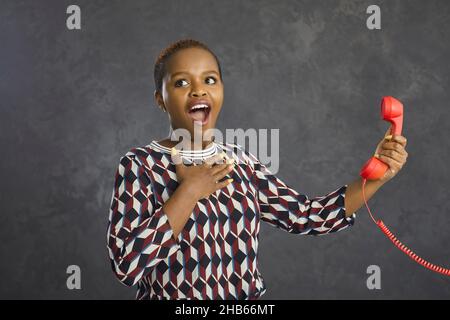 Dunkelhäutig schockierte Frau mit rotem retro Telefonhörer mit Draht auf grauem Hintergrund. Stockfoto