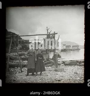 Magische Laternenrutsche von Mutter und Tochter stehend durch Holzrahmen von Kabeljau Fisch Trocknen mit Dorf im Hintergrund, Norwegen, c 1900 Stockfoto