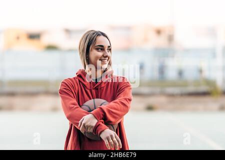 Blonde junge Frau schaut zur Seite, während sie einen Basketball im Freien hält Stockfoto