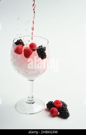 Gießen Sie Himbeere und Brombeere Gin in ein leeres Ballonglas mit Himbeeren und Brombeeren darin auf weißem Hintergrund Stockfoto