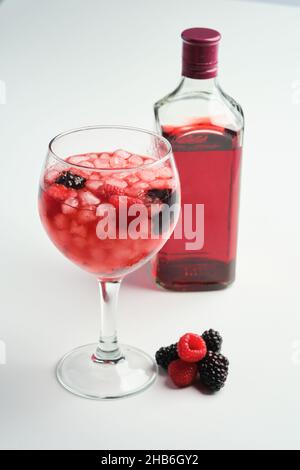 Ballonglas voller Eis und Himbeeren und Brombeeren Gin mit einer Flasche roten Gin auf einem weißen Background Stockfoto