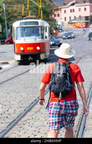 Tourist mit rotem T-Shirt und weißem Hut von der Rückseite zu Fuß in der Hauptstraße in Richtung der öffentlichen Straßenbahn. Prag. Reise- und Wandertouren auf Tschechisch RE Stockfoto