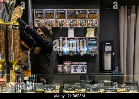 Eine Verkäuferin greift nach einem Beutel gerösteten Kaffees auf dem Regal eines Kaffeestandes in der Markthalle von Narbonne, Département Aude, Frankreich. Beschriftete Spender mit Rohkaffeebohnen aus Ländern wie Nicaragua, Kolumbien, Guatemala, Äthiopien, Peru und Indien säumen die Wand hinter ihr. Stockfoto