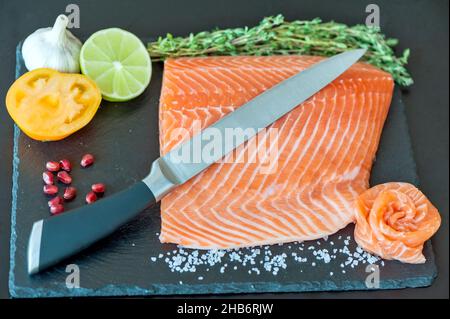 Koch mit Messer rohen Lachs auf Hackblock schneiden. Lachsfiletsteak und Gewürze auf schwarzem Schneidebrett. Stockfoto