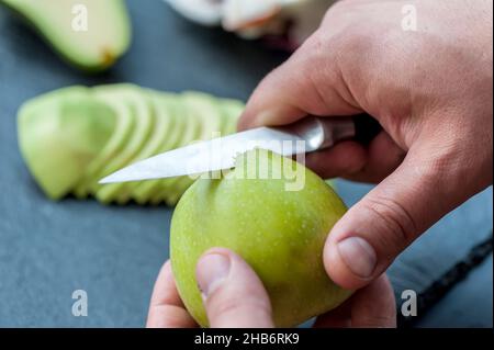 Hand mit Messer, das einen Apfelkern auf einem dunklen Schneidebrett ausschneidet, frischer saftiger grüner Apfel, der auf dem schwarzen Hintergrund gestreut ist Stockfoto