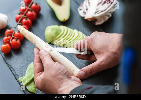 Handschälende Spargelstämme mit Messer, entfernen äußere Blätter, stapelbeschnittener Spargel auf dunkler Oberfläche liegend Stockfoto