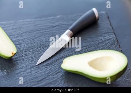 Männliche Hände, die die frische Avocado mit einem Küchenmesser des Küchenchefs auf einem mattschwarzen Schneidebrett schneiden. Gesundes Essen und Kochkonzept Stockfoto