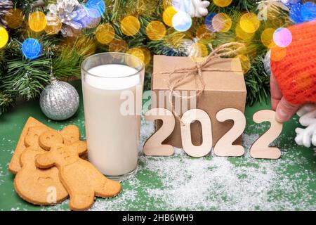 Kekse, ein Glas Milch für den Weihnachtsmann auf einem mit Schnee bedeckten Tisch auf dem Hintergrund von Weihnachtsdekorationen und Geschenken. Hand hält Holznummer 2 Zoll Stockfoto