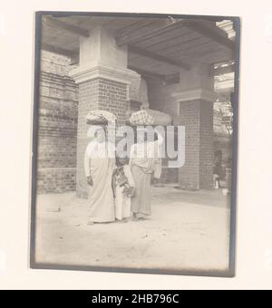 Ein Mann und eine Frau mit Gegenständen auf dem Kopf und ein Kind dazwischen in der Nähe einer Buddha-Statue, Südostasien, Jan Adriani (zugeschrieben), Südostasien, 1907, fotografische Unterstützung, Silbergelatine-Druck, Höhe 114 mm × Breite 89 mm Stockfoto