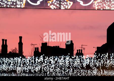 Weihnachtseinkäufe im Stadtzentrum Lichter mit Kaminen und Geschäfte, die bei Sonnenuntergang silhouettiert wurden. Schöner rosafarbener Winterhimmel mit fernsehantennen und Straßenbeleuchtung Stockfoto