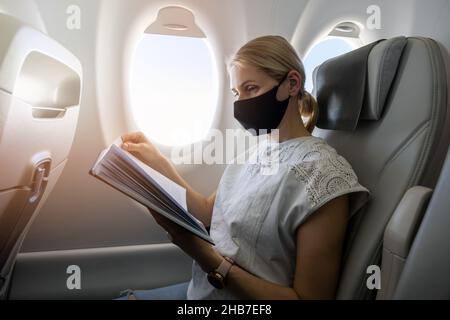 Frau trägt Gesichtsmaske und Lesebuch im Flugzeug. Reise in der Pandemie Stockfoto