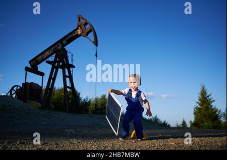 Junge Kind Ingenieur Spaß im Freien mit Ölpumpenheber an sonnigen Sommertag. Lächelndes Kind mit kleinem Solarmodul. Alternative fotovoltaische Ökoenergiekonzeption. Stockfoto