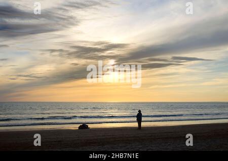 Ein Rentner im Hut geht bei Sonnenuntergang am Meer entlang der Uferpromenade Stockfoto