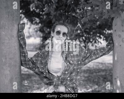 Zwischen den Säulen im Park steht eine süße Dame in Sonnenbrille mit zwei Zöpfen im karierten Hemd mit sehr langen Ärmeln. Schwarzweiß-Foto Stockfoto