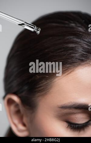 Nahaufnahme einer jungen Frau, die eine Pipette mit Öl in der Nähe von glänzendem Haar hält, das auf Grau isoliert ist Stockfoto