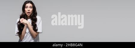 Überraschte Frau mit langen welligen Haaren und geballten Händen isoliert auf grauem Banner Stockfoto
