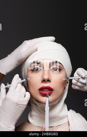 Plastische Chirurgen in Latexhandschuhen, die ein Operationsmesser und Spritzen in der Nähe der Frau in einem elastischen Verband auf dem Kopf halten, isoliert auf dunkelgrau Stockfoto